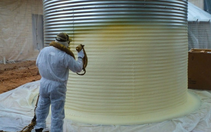  A worker in protective gear sprays insulation foam onto the exterior of a large metal tank, with protective sheets covering the ground. Insulation services for roofs, water and sewage tanks from ayahkutub.com.