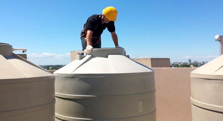 A worker wearing a yellow hard hat and gloves cleaning a large rooftop water tank under a clear blue sky. Cleaning water tanks and sewage tanks from ayahkutub.com.